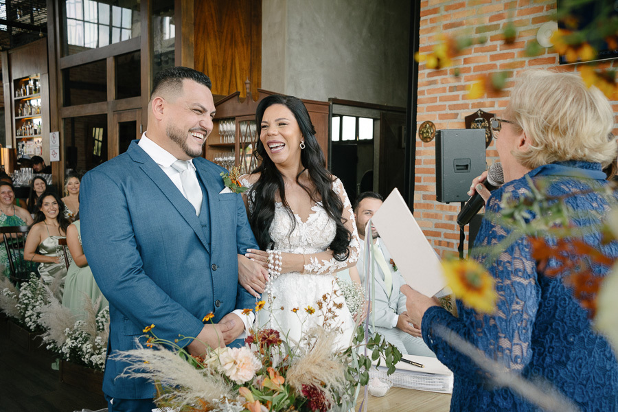 Os noivos Letícia e Juliano dão gargalhadas contagiantes no altar durante a celebração no Troina. A noiva segura um terço enquanto a celebrante fala ao microfone. O momento irradia a felicidade e animação sem igual desse casamento em São José dos Campos.