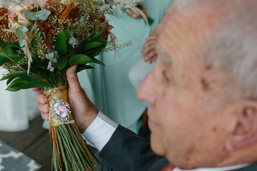 Close-up das mãos do pai da noiva segurando o buquê colorido, destacando um relicário prateado com a foto de duas mulheres preso ao caule. A imagem simboliza a memória e o amor familiar presentes na cerimônia de Letícia e Juliano no Troina.