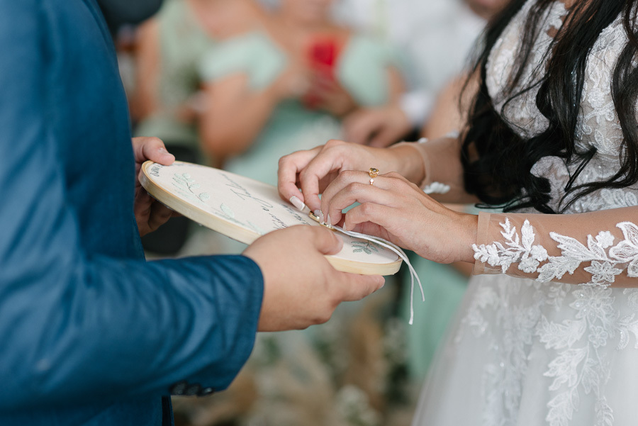 Close-up das mãos de Letícia e Juliano durante a troca de alianças no altar do Troina. O noivo segura um bastidor bordado que serve como porta-alianças, enquanto a noiva retira as joias. O momento simboliza a união e o amor do casal em São José dos Campos