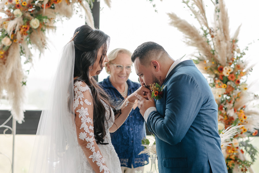 O noivo Juliano beija ternamente a mão da noiva Letícia durante a cerimônia no Troina. Ela veste um vestido rendado com véu longo, enquanto ele usa terno azul. Ao fundo, a celebrante sorri diante de uma luxuosa decoração com flores e capim-dos-pampas.