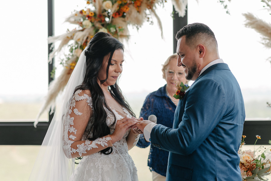 Noiva Letícia coloca a aliança no dedo do noivo Juliano durante a cerimônia no Troina. Ela está de perfil, com véu longo, enquanto ele sorri emocionado. Ao fundo, a celebrante observa o momento diante de um arco de flores secas e naturais.