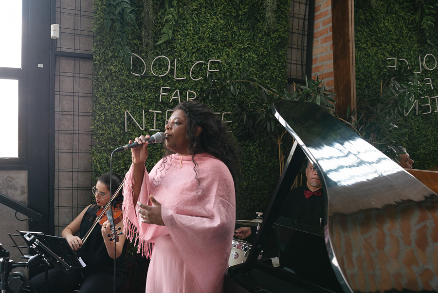 Cecília Militão solta a voz ao lado de uma violinista e um pianista durante o casamento no Troina. Ao fundo, uma parede de folhagens com o letreiro neon 
