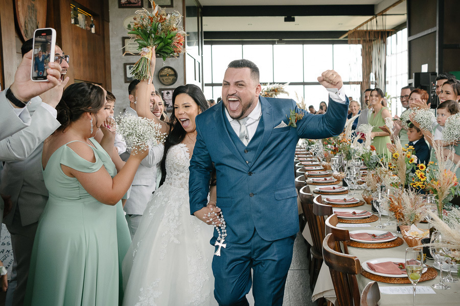 Os noivos Letícia e Juliano comemoram com euforia a saída da cerimônia no Troina. Juliano vibra com o punho erguido, enquanto Letícia levanta seu buquê. Convidados celebram ao redor de uma longa mesa posta, em um momento de alegria contagiante no casament