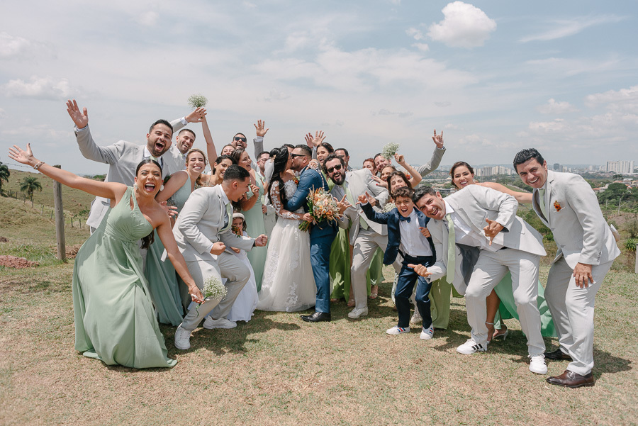 Letícia e Juliano se beijam cercados por padrinhos e madrinhas em um gramado ensolarado. O grupo celebra com euforia, braços erguidos e sorrisos contagiantes, capturando a essência da festa inesquecível e a animação sem igual no casamento no Troina.