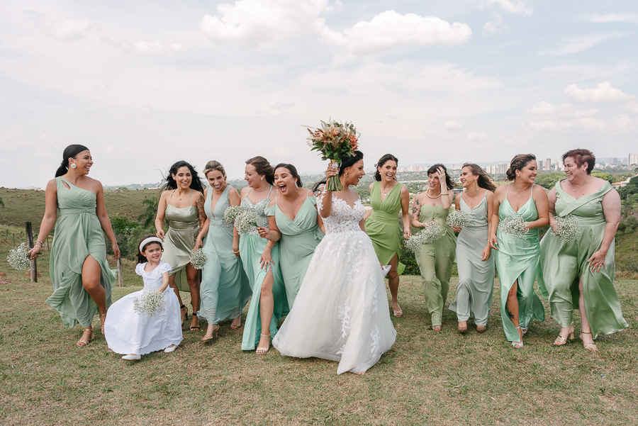 A noiva Letícia caminha alegremente em um gramado, erguendo seu buquê, acompanhada por suas madrinhas vestidas em tons de verde. O grupo sorri e celebra com animação sob um céu claro, capturando a felicidade contagiante do casamento no Troina.