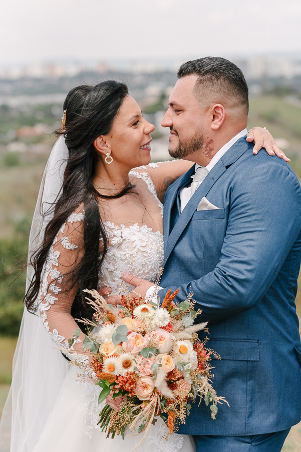Os noivos Letícia e Juliano trocam olhares apaixonados em um abraço ao ar livre em São José dos Campos. Letícia sorri segurando o buquê colorido, enquanto Juliano a envolve com carinho, eternizando a intensidade do amor do casal após o sim no Troina.