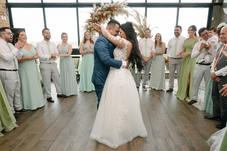 Os noivos Letícia e Juliano dançam abraçados no centro do salão do Troina, cercados por seus padrinhos e madrinhas que aplaudem sorridentes. O casal compartilha um momento íntimo e romântico, celebrando o início da festa em São José dos Campos.