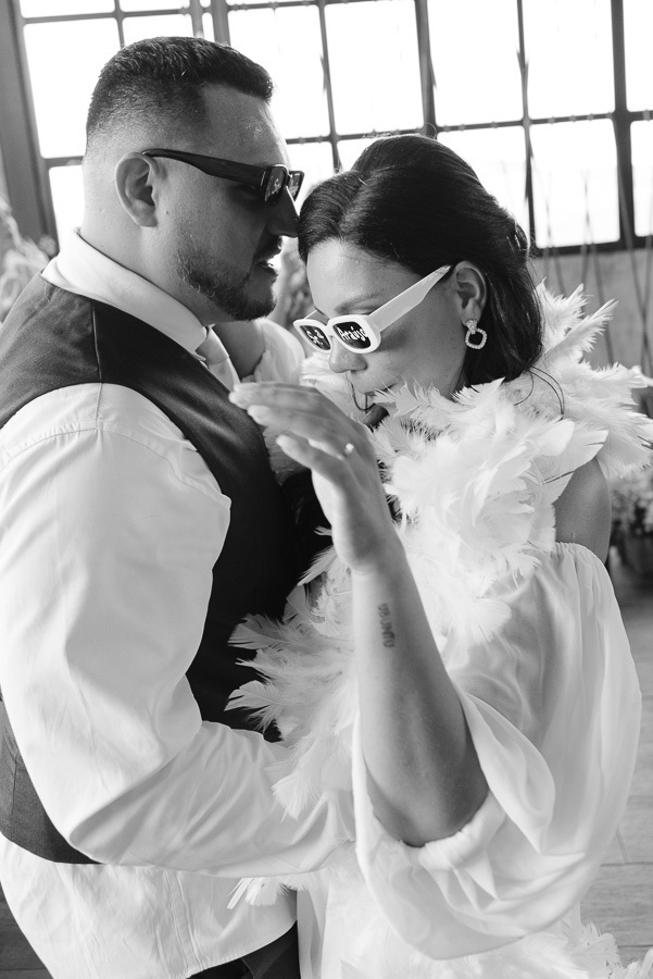 Foto em preto e branco dos noivos Letícia e Juliano em um momento descontraído na festa. Ambos usam óculos escuros, e a noiva complementa seu segundo look com uma estola de plumas branca, celebrando com muita personalidade e estilo no Troina.