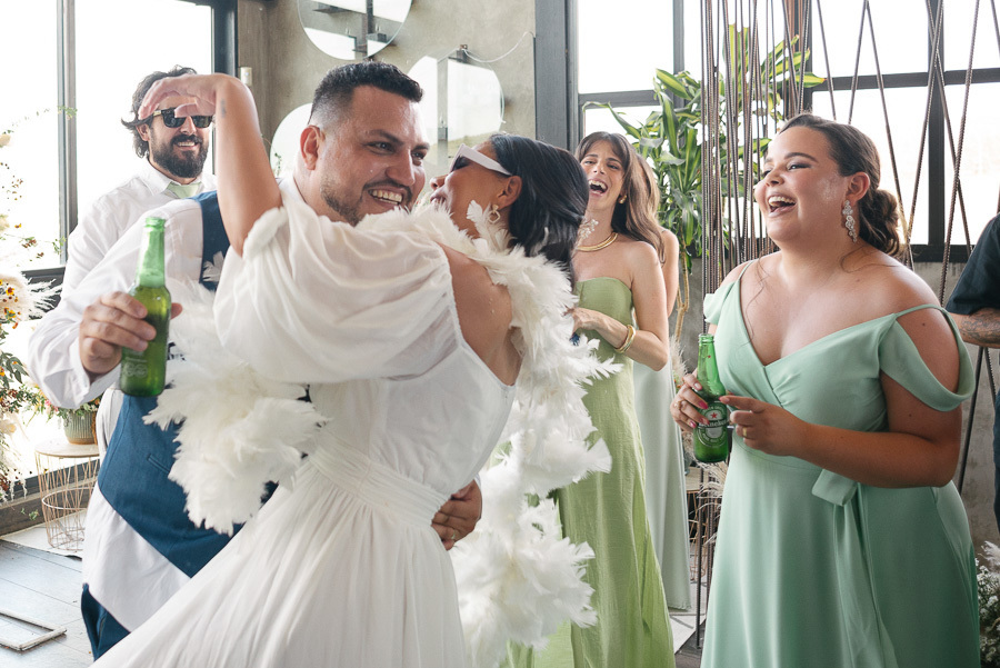 Os noivos Letícia e Juliano dançam e brindam com cerveja na pista do Troina. Ela usa o segundo look com plumas e óculos escuros, enquanto ele sorri abraçado à esposa. Madrinhas de verde celebram ao lado, em um momento de pura animação e descontração.
