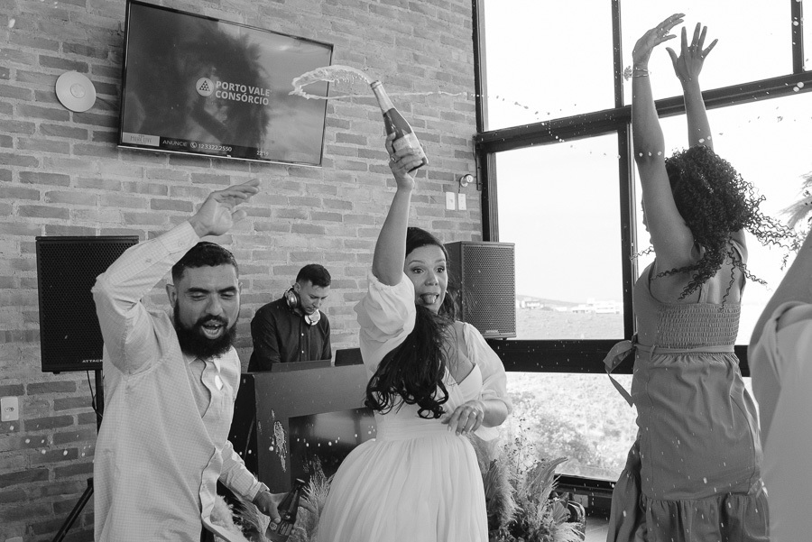 Noivos Letícia e Juliano dançam com empolgação na pista do Troina. Ela usa seu segundo vestido branco fluido e ele está de colete. Ao redor, convidados celebram com braços erguidos, capturando a energia e a alegria contagiante da festa em São José.