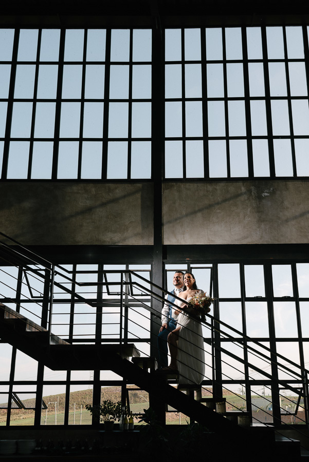 Silhueta dos noivos Letícia e Juliano em uma escada moderna no Troina. Eles posam diante de uma imensa parede de vidro que revela a paisagem de São José dos Campos, criando um jogo de luz e sombra que encerra com elegância o registro do casamento.