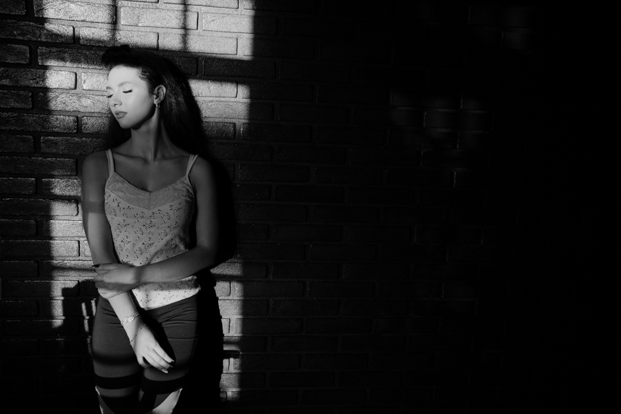 Um retrato artístico e dramático em preto e branco. O jogo de luz e sombras na parede de tijolos realça a beleza da jovem durante um momento de pausa no ensaio.