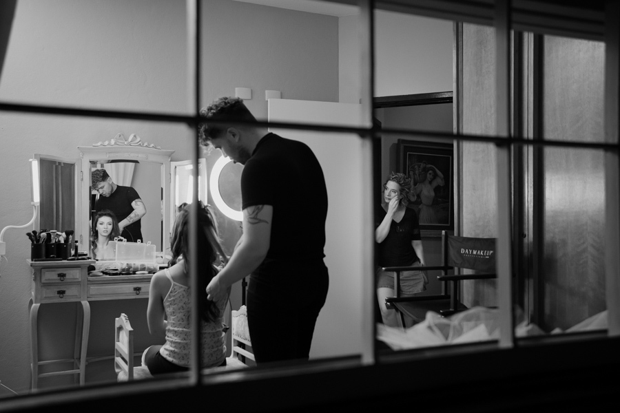 Bastidores de um sonho em preto e branco. Uma visão documental e ampla do ambiente de making-of, mostrando a equipe e a debutante se preparando para a festa.