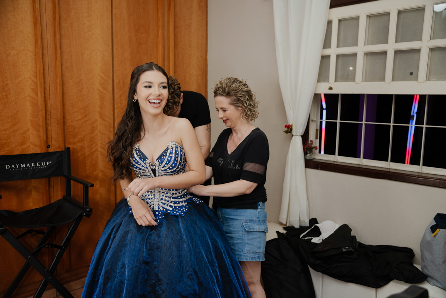 Risadas e muita emoção nos bastidores. O momento divertido de vestir o deslumbrante vestido de valsa azul, com ajuda especial, durante a preparação para os 15 anos.