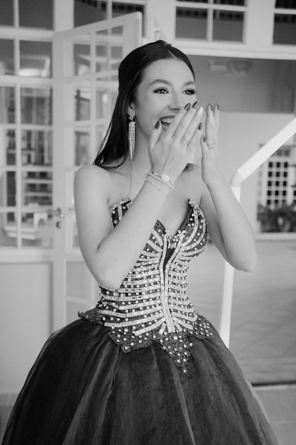 Emoção à flor da pele. Em preto e branco, a debutante cobre o rosto em um gesto delicado de surpresa e felicidade durante os preparativos de sua festa.
