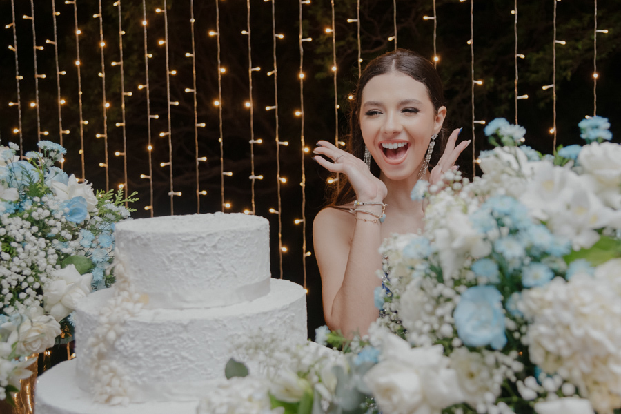 A emoção do grande dia! Debutante encantada ao lado do bolo e da decoração floral azul e branca. No Buffet Dona Lucy em Jacareí - SP