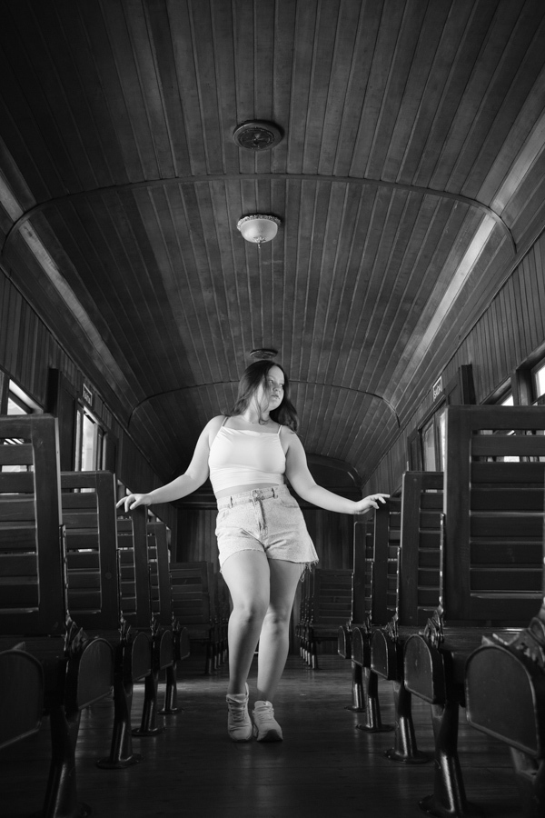 Simetria no vagão: Kiara em pé no centro do vagão de passageiros. Fotografia artística explorando as linhas e luz da Maria Fumaça.