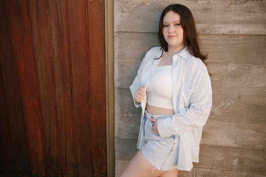 Look casual: Kiara com short jeans e camisa branca em frente à madeira rústica. Fotos descontraídas e joviais.