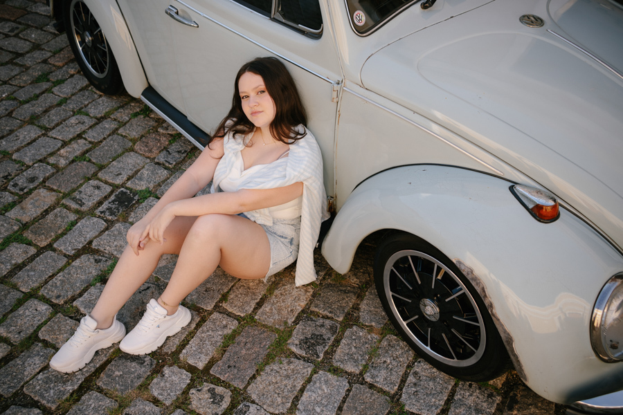 Vibe retrô divertida: Kiara sentada no paralelepípedo ao lado de um Fusca branco. Ensaio externo urbano e cheio de personalidade.