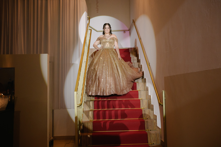 Entrada de gala. A debutante posa com vestido dourado na escadaria com tapete vermelho, valorizando a arquitetura do Espaço Lisbon em Jacareí.