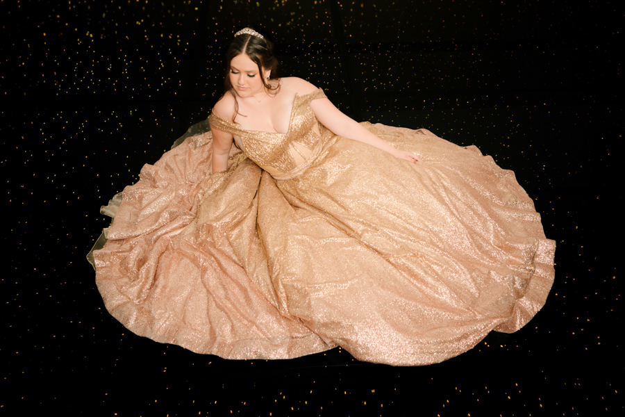 Pose de conto de fadas. Debutante sentada no chão com o vestido dourado espalhado, criando uma imagem mágica em Jacareí.