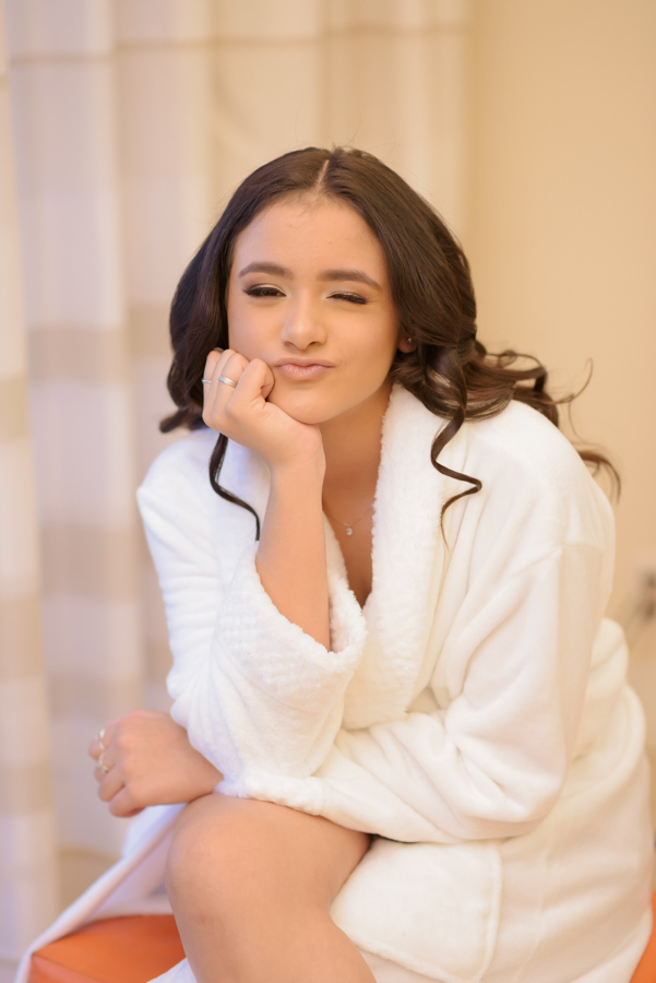 Fotografia de 15 anos da atriz Juju Salini no making of no Espaço Le Six, em Jacareí. Juju usa roupão branco, pisca um olho e faz pose descontraída enquanto se prepara para a festa de debutante. Um momento leve e espontâneo capturado pelo fotógrafo.