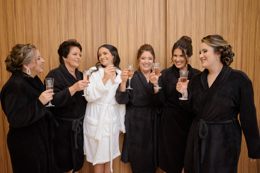 Brinde no making of de 15 anos no Espaço Le Six, Jacareí. A debutante Juju Salini em robe branco celebra com amigas em robes pretos, segurando taças de champanhe em um momento de descontração e alegria registrado pelo fotógrafo de debutantes.