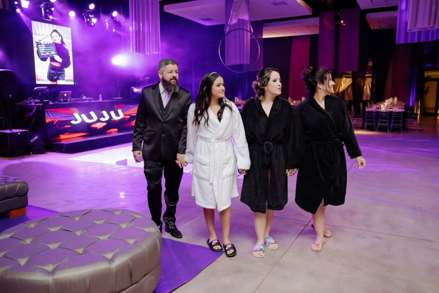 Juju Salini caminha pelo salão do Espaço Le Six em Jacareí durante o ensaio de 15 anos. Debutante de roupão branco de mãos dadas com família em cenário com iluminação cênica roxa e painel de LED ao fundo. Foto de bastidores da festa.
