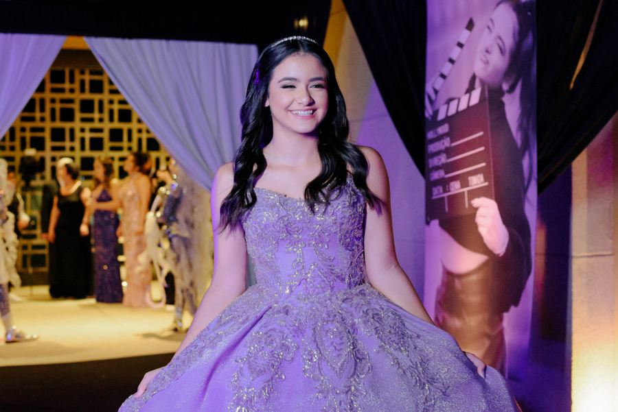 Debutante Juju Salini sorridente com vestido lilás de gala brilhante no Espaço Le Six, em Jacareí. Ao fundo, decoração temática de cinema e convidados. Fotografia de 15 anos profissional capturando a elegância e alegria da aniversariante.