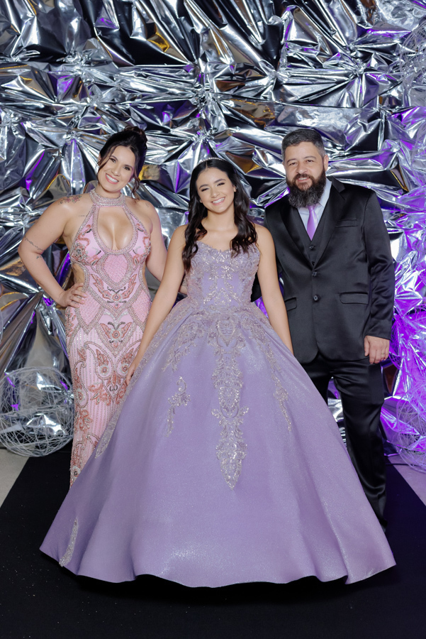 Retrato de família no aniversário de 15 anos de Juju Salini. A debutante posa com seus pais diante de um painel decorativo metalizado no Espaço Le Six, em Jacareí. Registro clássico e elegante da celebração de debutante.