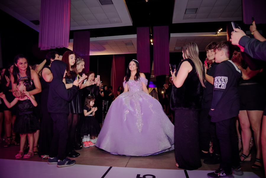 Debutante Juju Salini entra no salão principal do Espaço Le Six, em Jacareí, sob aplausos dos convidados. Ela usa um deslumbrante vestido de gala lilás com brilhos, sorrindo enquanto caminha pela pista em um momento emocionante da sua festa de 15 anos.