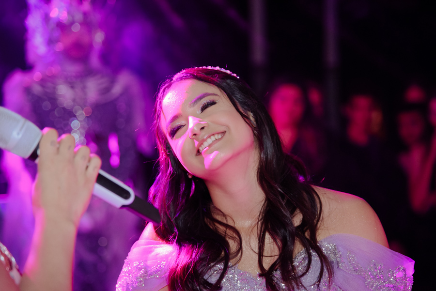 Juju Salini sorri emocionada durante as homenagens em sua festa de 15 anos no Espaço Le Six, em Jacareí. A debutante usa seu vestido de gala lilás sob iluminação cênica vibrante, enquanto ouve atentamente ao microfone em um momento de pura felicidade.
