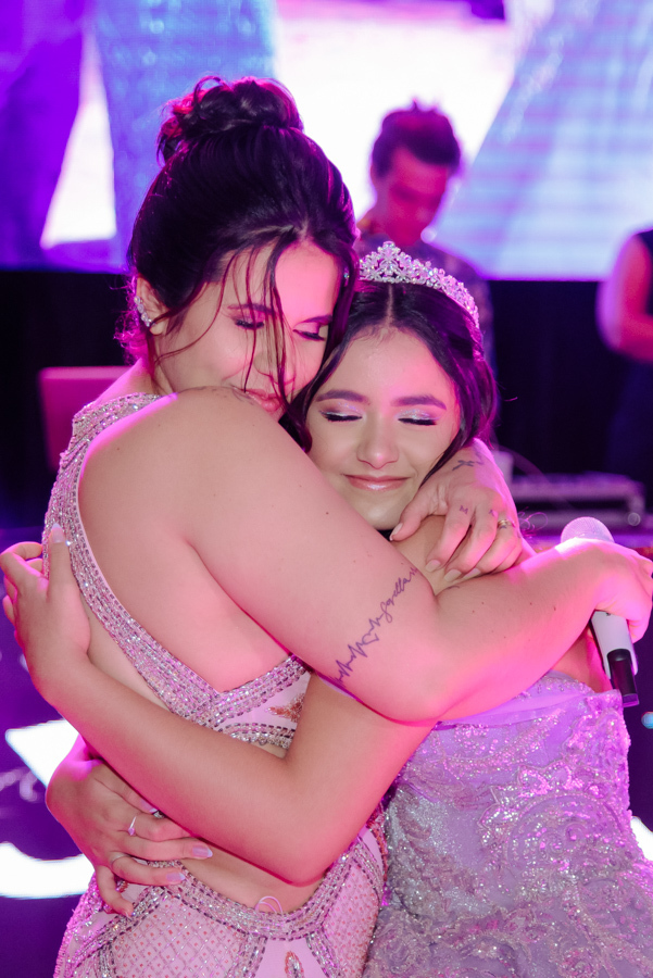 Juju Salini, com seu vestido de gala lilás e tiara, compartilha um abraço emocionado e carinhoso durante as homenagens em sua festa de 15 anos no Espaço Le Six, em Jacareí. O registro captura a conexão e a emoção de um dos momentos mais marcantes da noite