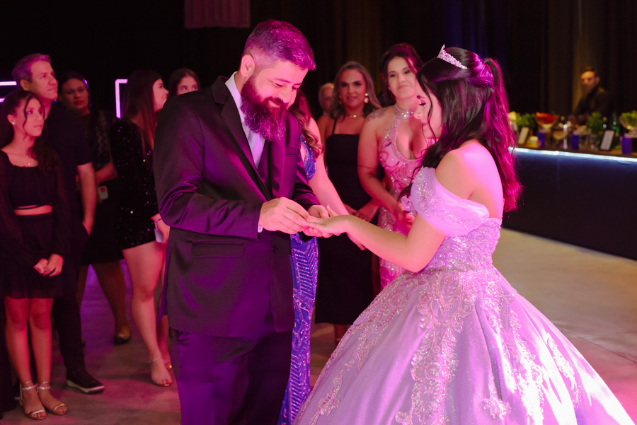 Momento solene da troca de anel entre a debutante Juju Salini e seu pai durante o protocolo de 15 anos no Espaço Le Six, em Jacareí. O registro captura a emoção da família e o detalhe do vestido de gala lilás sob a luz vibrante da festa.