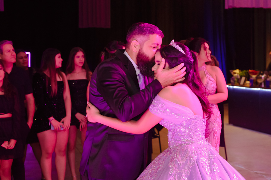 Momento emocionante entre a debutante Juju Salini e seu pai durante a festa de 15 anos no Espaço Le Six, em Jacareí. O pai abraça e beija a testa da filha com carinho, celebrando esse marco especial. Registro afetivo sob a iluminação vibrante do evento.