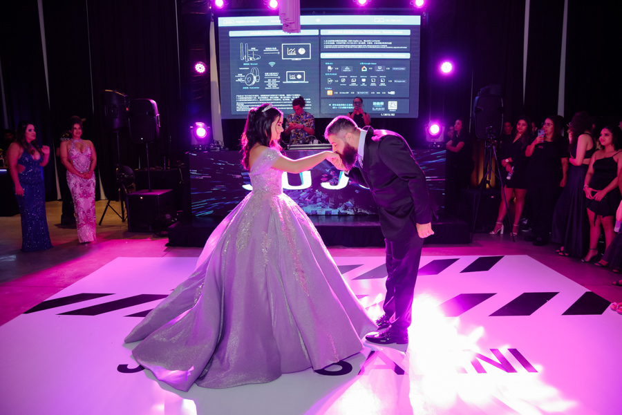 Momento solene da valsa de 15 anos no Espaço Le Six, em Jacareí, onde o pai beija carinhosamente a mão da debutante Juju Salini na pista de dança personalizada. O registro destaca a amplitude do salão, a iluminação vibrante e o deslumbrante vestido lilás.