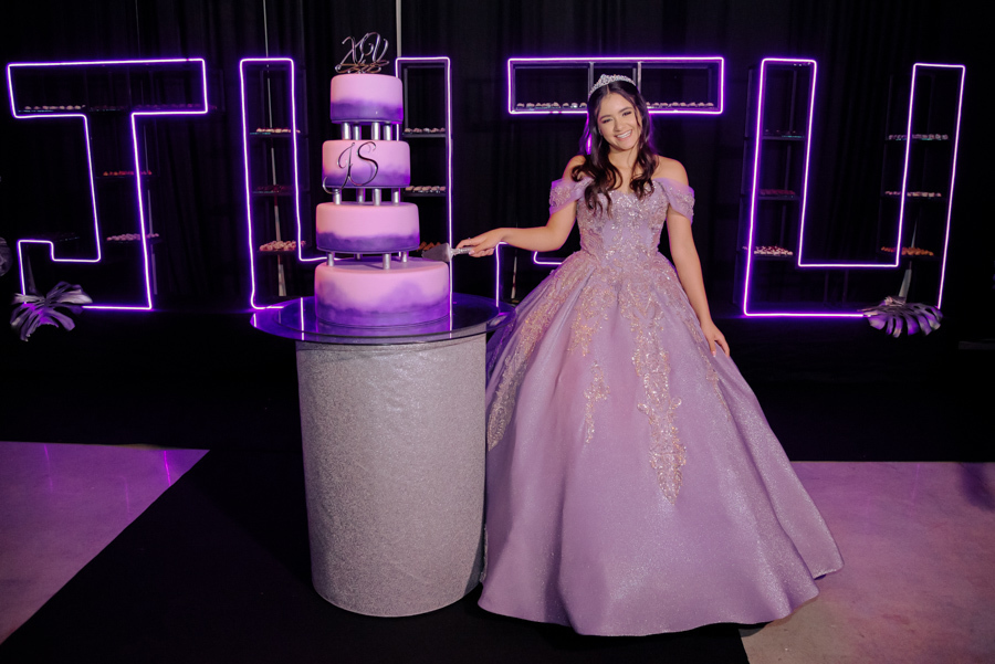 Juju Salini posa sorridente ao lado do seu bolo de 15 anos de quatro andares em tons de lilás e degradê no Espaço Le Six, em Jacareí. A debutante, com seu vestido de gala e tiara, destaca-se diante de uma moderna decoração com letreiro em neon personaliza