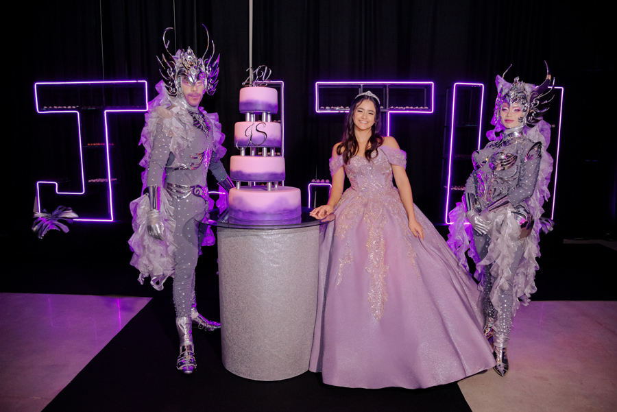 Juju Salini posa sorridente entre personagens lúdicos e futuristas ao lado de seu bolo de 15 anos no Espaço Le Six, em Jacareí. A debutante, em seu traje de gala lilás, completa o cenário moderno composto por letreiros em neon e uma decoração sofisticada.