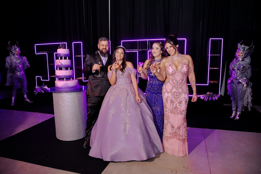 Brinde de celebração da debutante Juju Salini com sua família ao lado do bolo de 15 anos no Espaço Le Six, em Jacareí. O grupo sorri com taças em mãos, destacando o cenário moderno com letreiros em neon 
