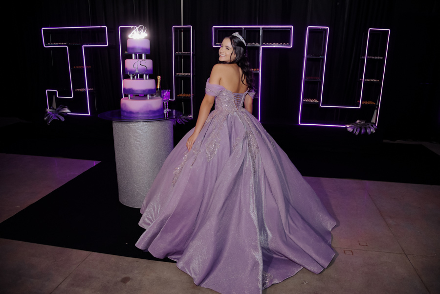 Juju Salini posa de costas exibindo os detalhes do espartilho de seu vestido de gala lilás no Espaço Le Six, em Jacareí. A debutante olha sorridente sobre o ombro ao lado do bolo de 15 anos, com a decoração moderna e letreiros em neon ao fundo.