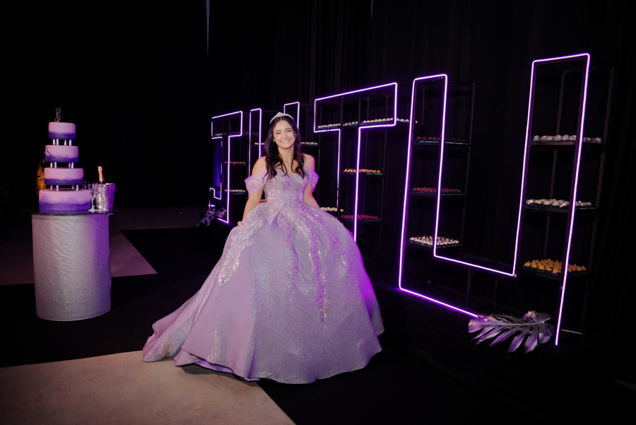 Juju Salini posa sorridente com seu vestido de gala lilás no Espaço Le Six, em Jacareí. Ao fundo, uma mesa de doces moderna com estantes iluminadas por LED roxo e um bolo de 15 anos sofisticado, completando o cenário luxuoso da festa de debutante.