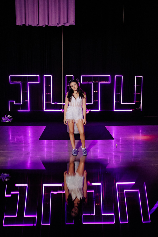 Retrato de Juju Salini em pé na pista de dança reflexiva do Espaço Le Six, em Jacareí. A debutante usa look moderno de franjas e tênis, posando diante do grande letreiro neon personalizado com seu nome para o ensaio da festa de 15 anos.