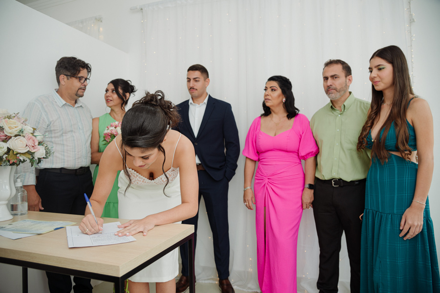 Noiva Karla assina documentos do casamento civil no Cartório de Jacareí sob o olhar de familiares e do noivo. Fotografia profissional para casamentos íntimos e cerimônias civis na região de SJC.