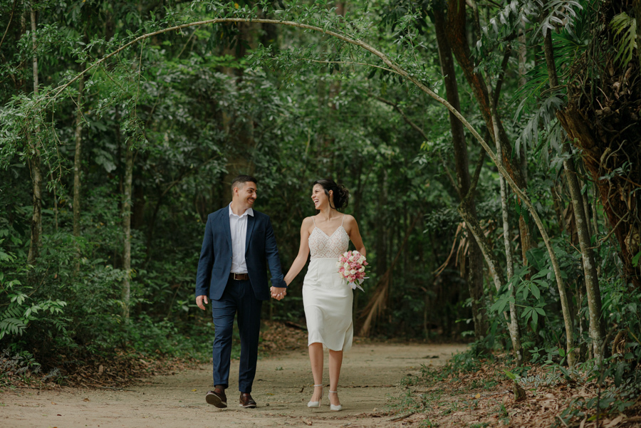 Noivos Karla e Diego caminham sorridentes em trilha arborizada durante ensaio no Parque da Cidade em São José dos Campos. Fotografia de casamento externa que destaca a leveza e a felicidade do casal após o casamento civil na região de SJC.