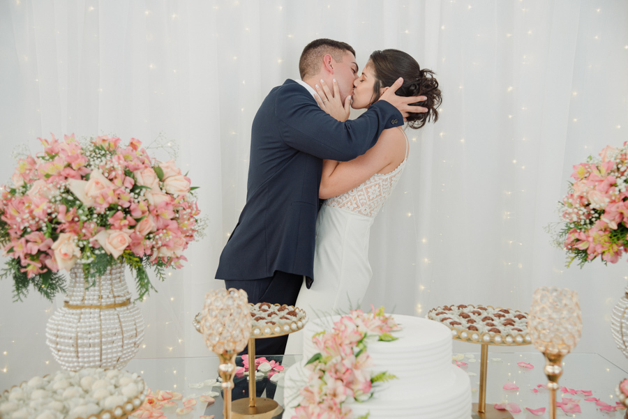 Noivos Karla e Diego se beijam atrás da mesa do bolo em recepção de casamento na chácara. Fotógrafo de casamento em Jacareí registra a comemoração e os detalhes da decoração romântica após a cerimônia civil.