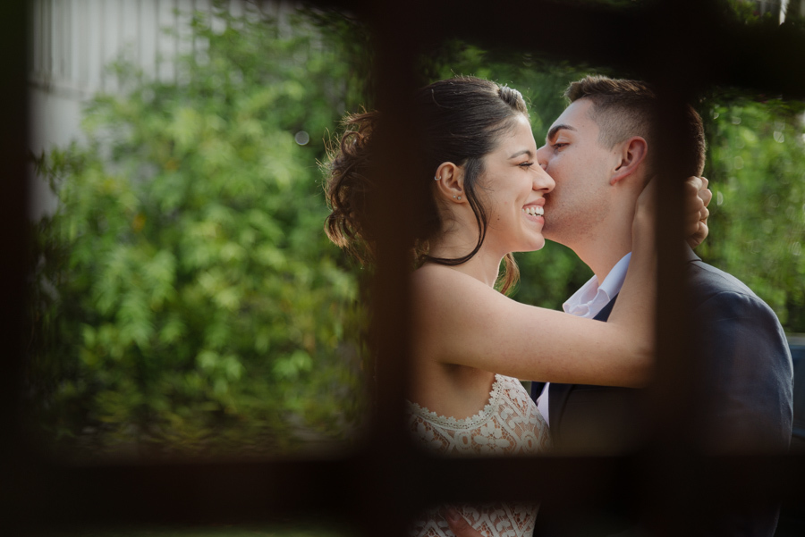 Noivo Diego beija o rosto da noiva Karla em momento de carinho durante a recepção. Fotógrafo de casamento em Jacareí utiliza técnica de composição criativa para registrar a felicidade e o sorriso da noiva após o casamento civil na região.