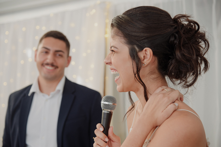 Noiva Karla sorri e segura microfone durante discurso emocionante na recepção, com o noivo Diego ao fundo. Fotógrafo de casamento em Jacareí registra momentos de alegria e espontaneidade na celebração após o casamento civil na região de SJC.