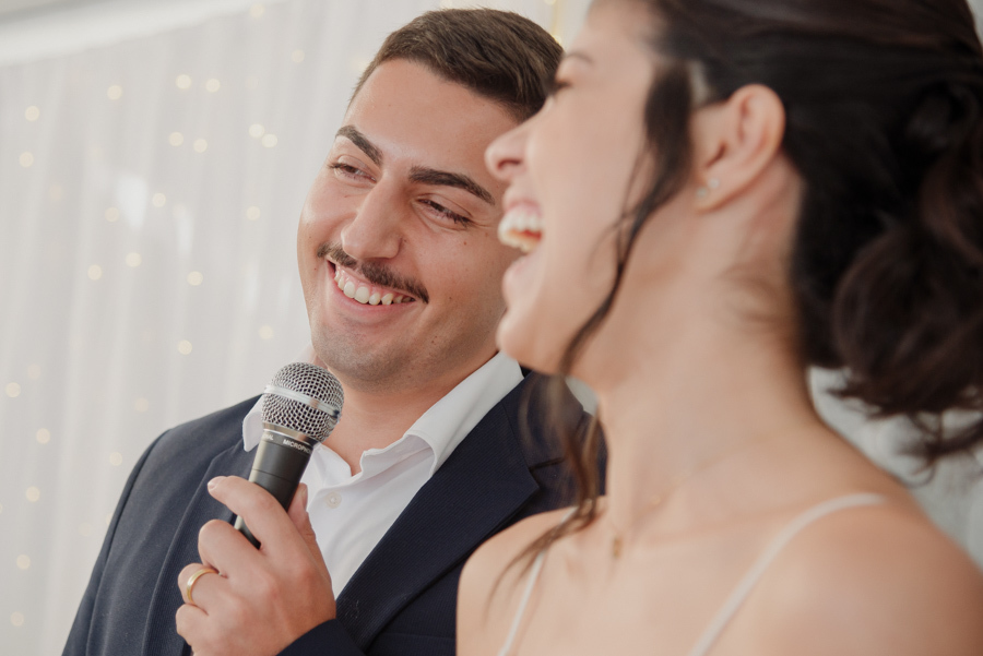 Close do noivo Diego sorrindo enquanto segura o microfone durante os votos na recepção. Fotógrafo de casamento em Jacareí captura a alegria e a espontaneidade do casal em celebração íntima após o casamento civil na região de SJC.