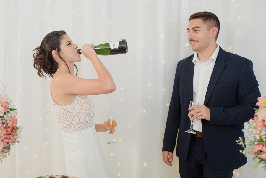 Noiva Karla bebe champanhe diretamente da garrafa em momento de descontração na recepção, com noivo Diego ao lado. Fotógrafo de casamento em Jacareí registra a alegria e celebração espontânea após o casamento civil no Vale do Paraíba.