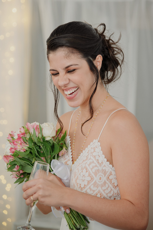 Noiva Karla sorri com alegria segurando seu buquê e uma taça de champanhe durante a recepção. Fotógrafo de casamento em Jacareí captura a felicidade e a espontaneidade da noiva após a cerimônia de casamento civil na região de SJC.